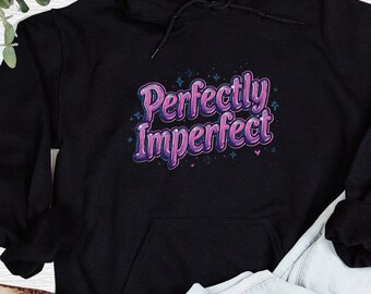 以下が含まれることがあります： 紫とピンクのグラデーションで「Perfectly Imperfect」の文字が入った黒いパーカー。パーカーにはフロントポケットとドローストリングフードが付いています。デザインには小さな星とハートのアクセントが含まれています。