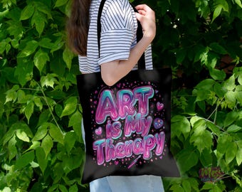 以下が含まれることがあります： 黒いトートバッグ。カラフルなバブルスタイルのフォントで「ART is MY Therapy」の文字がプリントされています。デザインにはハートと星が含まれています。バッグは緑の葉の背景の前にあります。