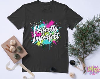 以下が含まれることがあります： ピンク、青、黄色のカラフルなペイントスプラッシュを背景に、白い文字で「Perfectly Imperfect」と書かれた黒いTシャツ。デザインには星のアクセントと滴るペイント効果が含まれています。Tシャツは平らな面に表示されています。