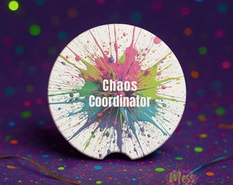 以下が含まれることがあります： ピンク、緑、青色のカラフルなペイントスプラッターデザインの白い車のコースター。「Chaos Coordinator」の文字が白で印刷されています。背景は、カラフルなボケのある濃い紫です。