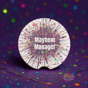 Könnte beinhalten: Weißer Auto-Untersetzer mit farbenfrohem Farbspritzer-Design. Der Text "Mayhem Manager" ist auf dem Untersetzer zentriert. Der Hintergrund ist ein dunkles Lila mit bunten Bokeh-Lichtern.