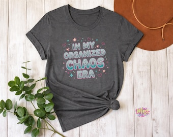 以下が含まれることがあります： 「IN MY ORGANIZED CHAOS ERA」の文字がキラキラとカラフルなデザインでプリントされたダークグレーのTシャツ。デザインには星、ハート、ダイヤモンドが含まれています。シャツは裾で結ばれています。