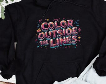以下が含まれることがあります： 黒色のフード付きスウェットシャツ。前面にポケットとドローコードが付いています。「COLOR OUTSIDE THE LINES」というフレーズが、カラフルなバブルスタイルの文字と装飾要素で表示されています。
