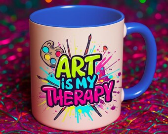 以下が含まれることがあります： 青いハンドルと内側を備えたベージュ色のセラミックマグ。マグカップには、鮮やかな色の「ART is MY THERAPY」という文字が、パレット、筆、ペイントスプラッシュとともに描かれています。楽しくて芸術的なデザインです。