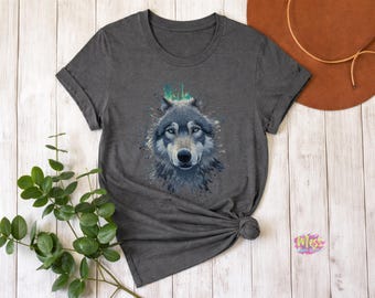 以下が含まれることがあります： 水彩画風のオオカミの肖像画が描かれた濃いグレーのTシャツ。オオカミは青い目を持ち、頭の上に緑と青の飛沫があります。Tシャツは裾で結ばれています。