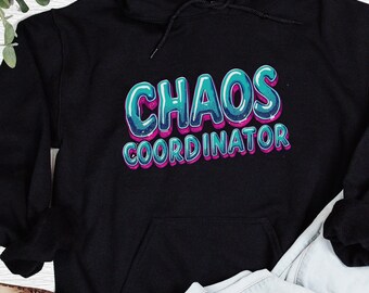 以下が含まれることがあります： 黒色のフード付きスウェットシャツ。文字「CHAOS COORDINATOR」が、ターコイズとピンクのバブルフォントでプリントされています。スウェットシャツにはフロントポケットとドローストリングフードが付いています。デザインは胸の中央に配置されています。