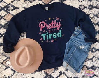 以下が含まれることがあります： ピンクとティール色の「Pretty But Tired」の文字がプリントされたネイビーブルーのスウェットシャツ。ハートと星が周りに描かれています。ベージュのフェドラハットとブルージーンズも写っています。カジュアルで快適なスタイルです。