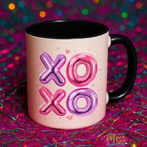 May include: A pink ceramic mug with a black handle and interior. The mug features the text "XO XO" in metallic purple and pink bubble letters, with small hearts and stars. The mug is set against a colourful, blurred background.