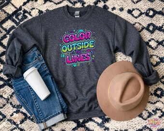 以下が含まれることがあります： カラフルなグラフィティスタイルのデザインで「COLOR OUTSIDE THE LINES」というテキストが入った濃いグレーのスウェットシャツ。画像には、青いジーンズ、白いコーヒーカップ、タン色のフェドラハットも含まれています。