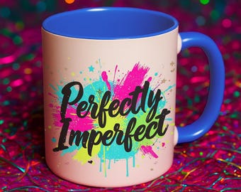 以下が含まれることがあります： 淡いピンク色の外側、青色の内側、青色のハンドルが付いたセラミックマグカップ。「Perfectly Imperfect」の文字が、カラフルなスプラッターデザインの上に黒い筆記体で書かれています。背景はカラフルな光のボケです。