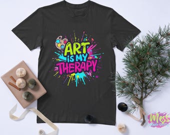 以下が含まれることがあります： 「ART is MY THERAPY」の文字がカラフルなグラフィティスタイルで描かれた黒いTシャツ。デザインには絵の具パレットとカラフルなペイントスプラッシュが含まれています。Tシャツは、装飾的な要素のある白い表面に表示されています。