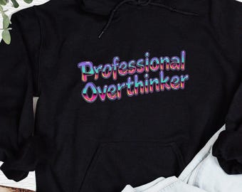 以下が含まれることがあります： カラフルなバブルフォントで「Professional Overthinker」と書かれた黒いフード付きスウェットシャツ。スウェットシャツにはフロントポケットとドローストリングフードが付いています。デザインは胸の中央に配置されています。