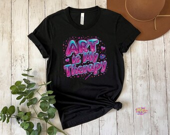 以下が含まれることがあります： 「ART is my Therapy」の文字が青とピンクのバブルレターでデザインされた黒いTシャツ。小さなハートと絵の具パレットのグラフィックが添えられています。Tシャツは裾で結ばれています。