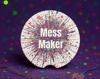 以下が含まれることがあります： 紫、ピンク、緑色のカラフルなペイントスプラッターデザインの白い車のコースター。「Mess Maker」の文字が中央にあります。背景は、カラフルなドットと「Mess Made Pretty」の文字が入った濃い紫です。