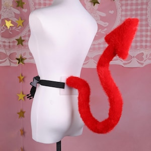 May include: A red devil tail accessory with a black adjustable belt. The tail is fluffy and shaped like an arrow at the end. The belt has a plastic buckle. The background is a light pink color.