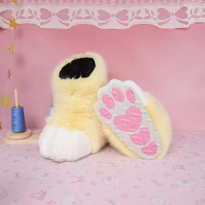 May include: A pair of plush, furry paw-shaped slippers. The slippers are primarily yellow with a black top, white toes, and a pink paw print design on the soles. The slippers are designed to resemble animal paws.
