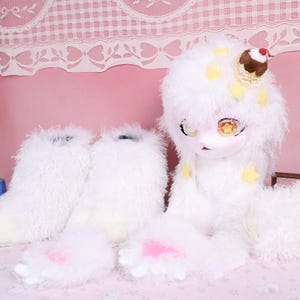 May include: A white, fluffy doll with matching boots and paw-like feet. The doll has a large head with a painted face, yellow star accents, and a small dessert on top. The boots and feet have pink accents.