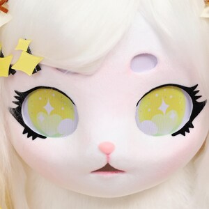 Puede incluir: Primer plano de una m&aacute;scara de animal de peluche blanco con ojos grandes y expresivos. Los ojos son amarillos con un dise&ntilde;o de estrella y nube, delineados en negro. La m&aacute;scara tiene una nariz y boca rosadas, con pelaje blanco y detalles de estrellas amarillas.