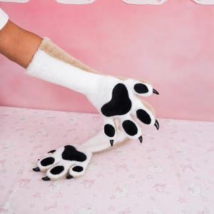 May include: A pair of long, plush animal paw gloves featuring a white and tan faux fur exterior. The palms are white with black velvet paw pads and sharp, glossy black claws attached to each of the four fingers on both gloves.