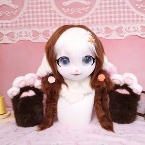 May include: A white and brown furry costume head with large blue eyes, long brown hair, and a white fur collar. The costume includes matching brown paw-shaped hand covers with pink pads. Decorative seashell accents are pinned to the costume.