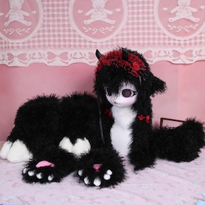 May include: A black and white plush doll with a cat-like appearance. The doll has a white face with large eyes, black fur, and a red headband with horns and roses. The doll's paws have pink pads.