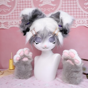 Puede incluir: Una cabeza de maniquí blanca con orejas de conejo grises y blancas, ojos en espiral y nariz rosa. Guantes de pata grises y blancos con almohadillas rosas a ambos lados del maniquí.