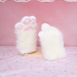 Puede incluir: Un par de guantes blancos y esponjosos con forma de pata y almohadillas rosas. Los guantes est&aacute;n hechos de suave piel sint&eacute;tica y est&aacute;n dise&ntilde;ados para parecerse a las patas de un gato. Se exhiben sobre un soporte de madera clara sobre un fondo rosa.