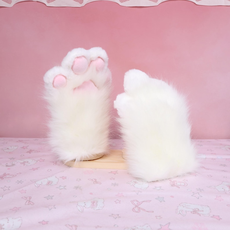 Puede incluir: Un par de guantes blancos y esponjosos con forma de pata y almohadillas rosas. Los guantes est&aacute;n hechos de suave piel sint&eacute;tica y est&aacute;n dise&ntilde;ados para parecerse a las patas de un gato. Se exhiben sobre un soporte de madera clara sobre un fondo rosa.