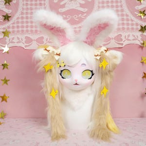 Puede incluir: Una m&aacute;scara blanca y rosa con orejas de conejo y detalles de estrellas amarillas. La m&aacute;scara tiene ojos grandes y expresivos y cabello rubio largo y suelto. Las orejas est&aacute;n forradas de rosa. Este disfraz es perfecto para cosplay o eventos tem&aacute;ticos.