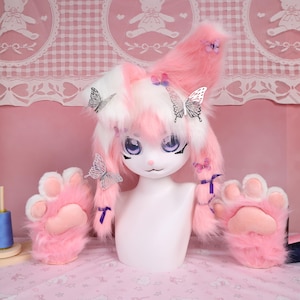 Può includere: Un set cosplay in pelliccia rosa e bianca con una testa di bambola con grandi occhi, orecchie soffici e guanti a forma di zampa. Le orecchie sono decorate con dettagli a farfalla e nastri viola. I guanti hanno un cuscinetto rosa.