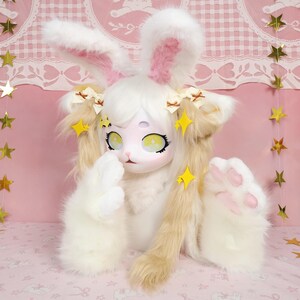 Puede incluir: Una mu&ntilde;eca con forma de conejo blanco y marr&oacute;n con orejas grandes, ojos amarillos y detalles en rosa. La mu&ntilde;eca tiene pelaje esponjoso y est&aacute; decorada con adornos amarillos en forma de estrella. Las patas de la mu&ntilde;eca tienen almohadillas rosas. El fondo es rosa con decoraciones de estrellas doradas.