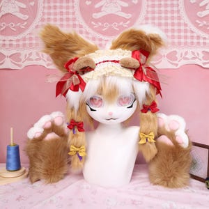 以下が含まれることがあります： 白い顔、ピンク色の目、茶色と白の毛皮の耳が特徴のウサギをテーマにしたコスチュームセット。セットには、足の形をした手袋、赤と黄色のリボン、レースのヘッドピースが含まれています。青い糸巻きも表示されています。