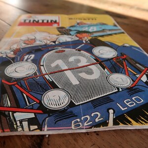 May include: A vintage comic book cover featuring a blue race car with the number 13 prominently displayed on its front grille. The illustration includes the text TINTIN in a red box at the top, along with the license plate number 622 L60.