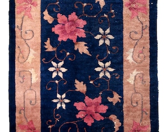 Op de afbeelding: Rechthoekig tapijt met een marineblauw midden en een bruinbeige rand. Het tapijt heeft bloempatronen in roze, wit en bruinbeige. Het bloemmotief omvat grote bloemen en kleinere bloesems met krullende ranken.