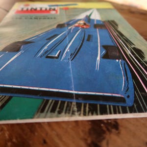 May include: Vintage comic book cover featuring a blue race car speeding down a track. The cover has the word "Tintin" at the top, with the name "J. Campbell" below. The artwork is in a retro style, with white and pink speed lines.