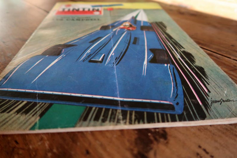 May include: Vintage comic book cover featuring a blue race car speeding down a track. The cover has the word "Tintin" at the top, with the name "J. Campbell" below. The artwork is in a retro style, with white and pink speed lines.