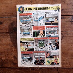 May include: A vintage comic book page titled "S.O.S. M&eacute;t&eacute;ores!" with multiple panels illustrating a snowy scene. The panels show cars, characters, and speech bubbles. The comic is displayed on a wooden surface.
