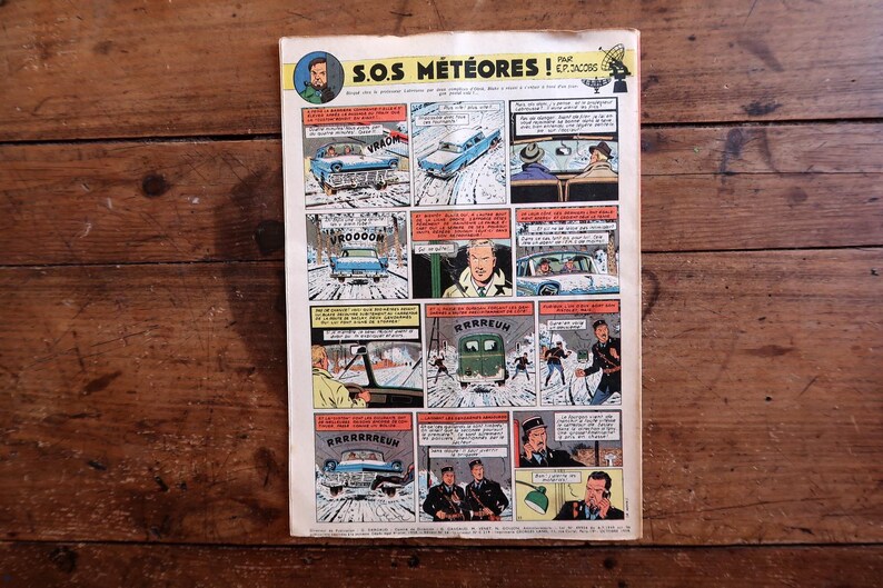 May include: A vintage comic book page titled "S.O.S. M&eacute;t&eacute;ores!" with multiple panels illustrating a snowy scene. The panels show cars, characters, and speech bubbles. The comic is displayed on a wooden surface.