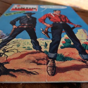 May include: Comic book cover featuring two cowboys in a desert landscape. One cowboy wears a black outfit and the other a red shirt and blue jeans. The comic book title is "Tintin" with text in French. The background shows cacti and mountains.