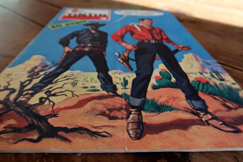 May include: Comic book cover featuring two cowboys in a desert landscape. One cowboy wears a black outfit and the other a red shirt and blue jeans. The comic book title is "Tintin" with text in French. The background shows cacti and mountains.