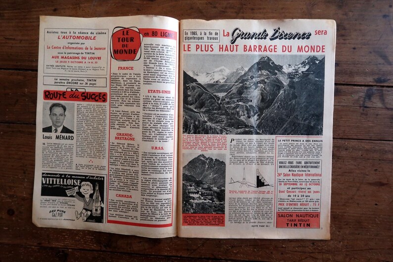 May include: An open vintage magazine with French text and illustrations. Headlines include "Le Tour du Monde" and "Le Plus Haut Barrage du Monde." The pages are on a wooden surface.