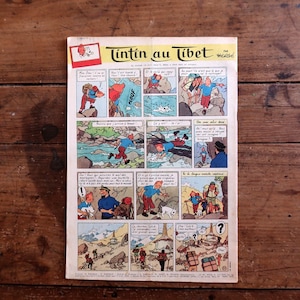 May include: A vintage comic book titled "Tintin au Tibet" in French, featuring colorful panels depicting adventure scenes. The cover has a red and white logo and is set against a wooden background.