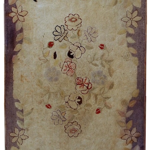 Op de afbeelding: Rechthoekig tapijt met een crèmekleurig middenveld, versierd met bloemmotieven in roze, rood en bruin. Het tapijt heeft een donkerpaarse rand en bloemmotieven langs de randen. Het ontwerp van het tapijt is symmetrisch.