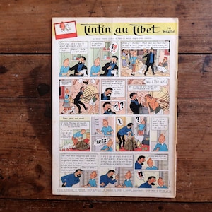May include: A vintage comic book titled "Tintin au Tibet" by Herg&eacute;, featuring panels with illustrations and speech bubbles. The cover is visible, set against a rustic wooden surface.