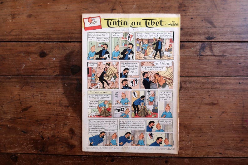 May include: A vintage comic book titled "Tintin au Tibet" by Herg&eacute;, featuring panels with illustrations and speech bubbles. The cover is visible, set against a rustic wooden surface.