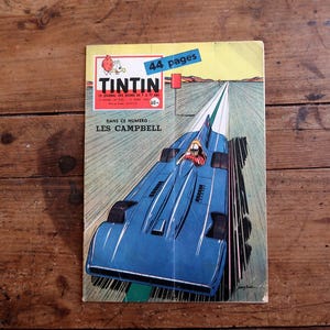 May include: A vintage comic book cover featuring a blue race car speeding down a track. The cover has the title "TINTIN" in red and white, with the text "44 pages" on a blue banner. The background is a wooden surface.