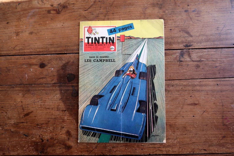 May include: A vintage comic book cover featuring a blue race car speeding down a track. The cover has the title "TINTIN" in red and white, with the text "44 pages" on a blue banner. The background is a wooden surface.
