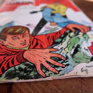 May include: A vintage comic book cover depicting a dramatic scene of a person in a red jacket reaching out, with another figure in blue and yellow. The artwork is in a classic comic book style, with bold colours and dynamic poses.