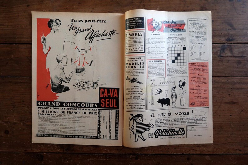May include: Open vintage magazine pages with illustrations and text in French. One page features a contest advertisement with figures and text, while the other has various ads, including stamps and a crossword puzzle. The pages are on a wooden surface.