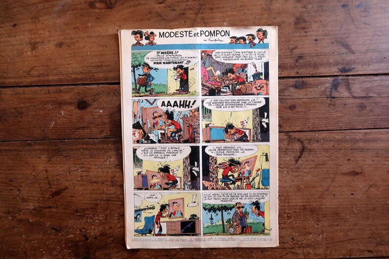 May include: A vintage comic book page titled Modeste et Pompon featuring a multi-panel grid with colorful illustrations and French text. The paper has visible aging, including yellowing edges and a central horizontal crease on the rectangular sheet.